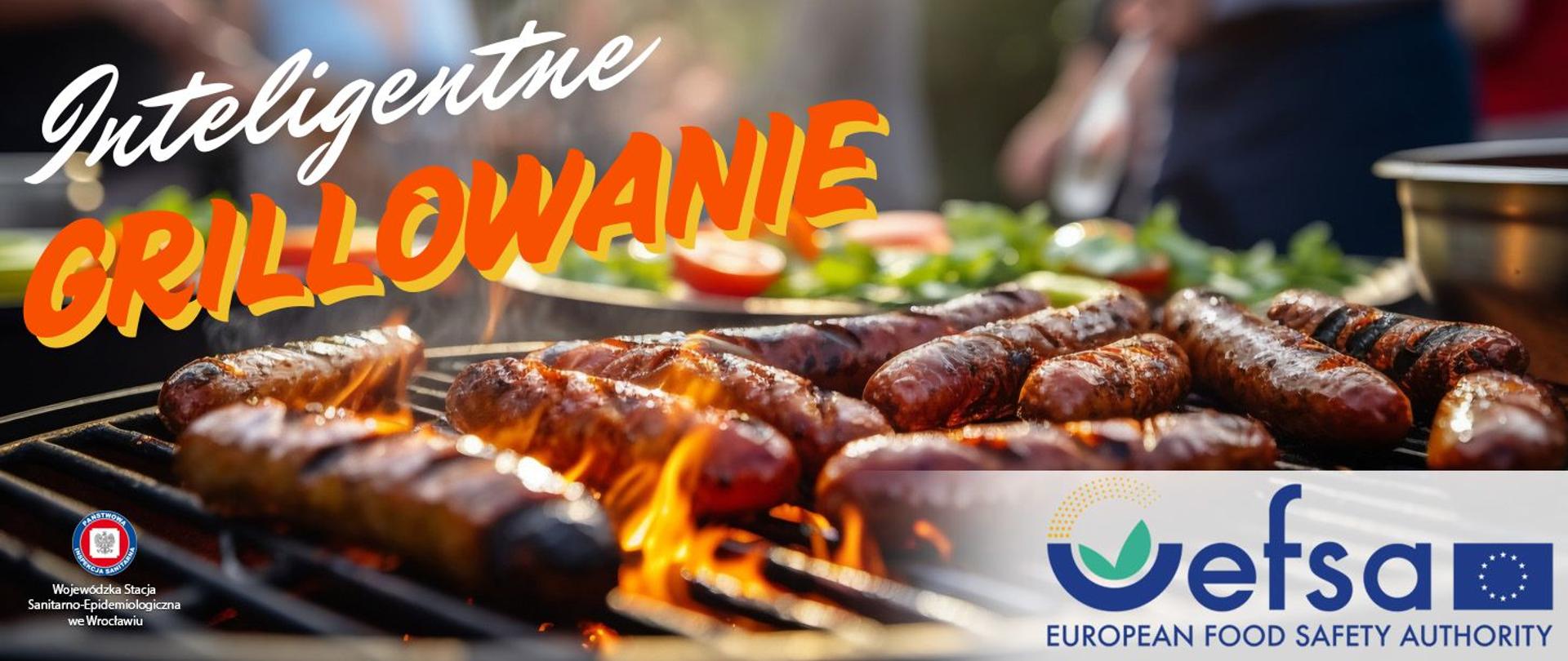 Kiełbaski piekące się na grillu, za nimi talerze z sałatą i pomidorami.