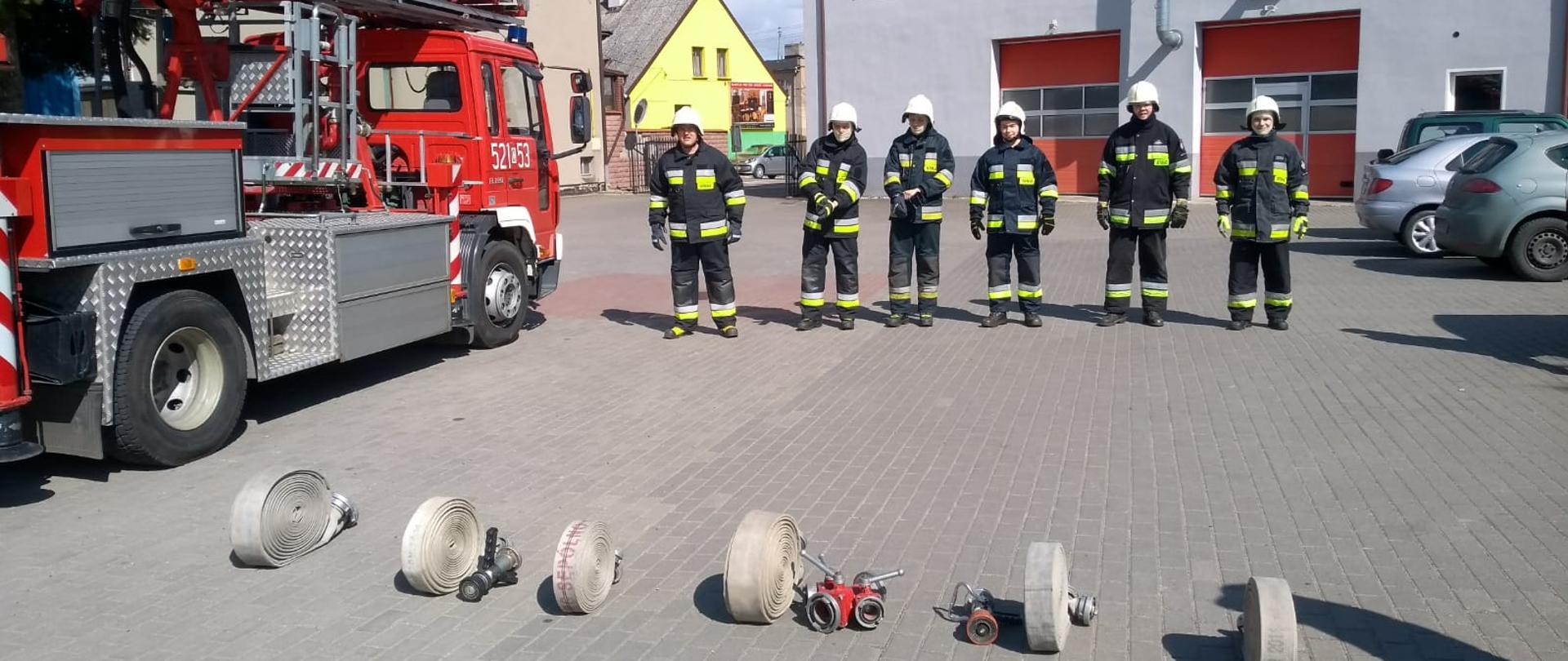 Zdjęcie wykonano w ciągu dnia na placu Komendy Powiatowej Państwowej Straży Pożarnej w Sępólnie Krajeńskim. Strażacy Ochotniczej Straży Pożarnej ubrani w mundury specjalne i hełmy przygotowują się do wykonania rozwinięcia bojowego. Na ziemi leżą odcinki węży tłocznych rozdzielacz strażacki oraz prądownice. Po lewej podnośnik hydrauliczny na podwoziu Volvo.