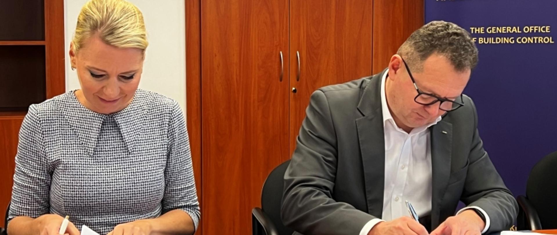 Dwie osoby podpisujących dokumenty przy stole; w tle baner „THE GENERAL OFFICE OF BUILDING CONTROL”.
