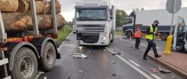 Na pasie ruchu stoi TiR który uderzył w ciężarówkę przewożącą bale drewniane. Ciężarówka zjechała z drogi do rowu. Z boku funkcjonariusz policji wykonujący czynności służbowe. 