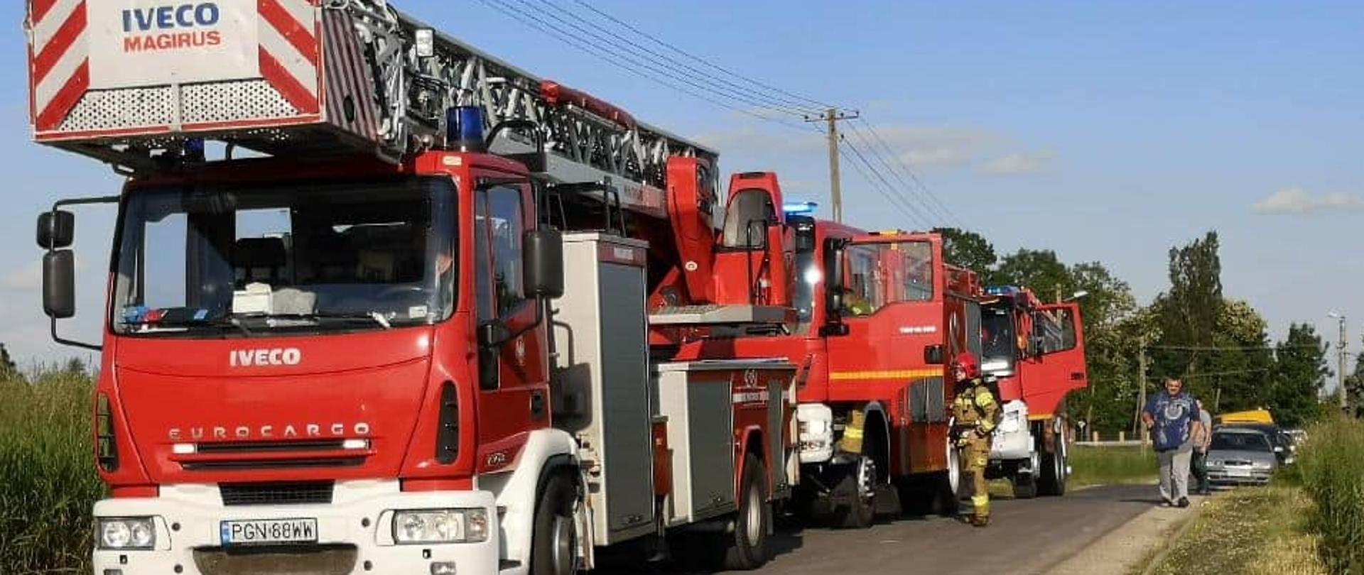 Słoneczny dzień. Trzy czerwone wozy strażackie stoją jeden za drugim na asfaltowej czarnej jezdni. W tle pole i drzewa zielone.