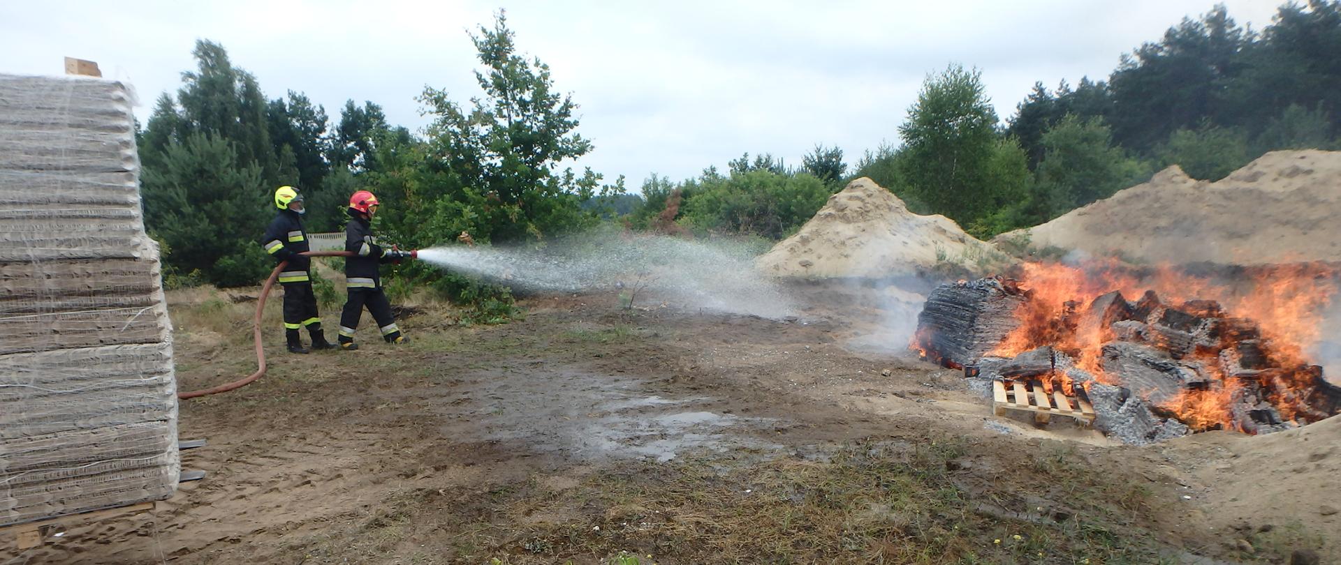 Dwóch strażaków trzyma wąż strażacki lejąc wodę przy składowisku palących się palet i wytłaczanek do jajek.