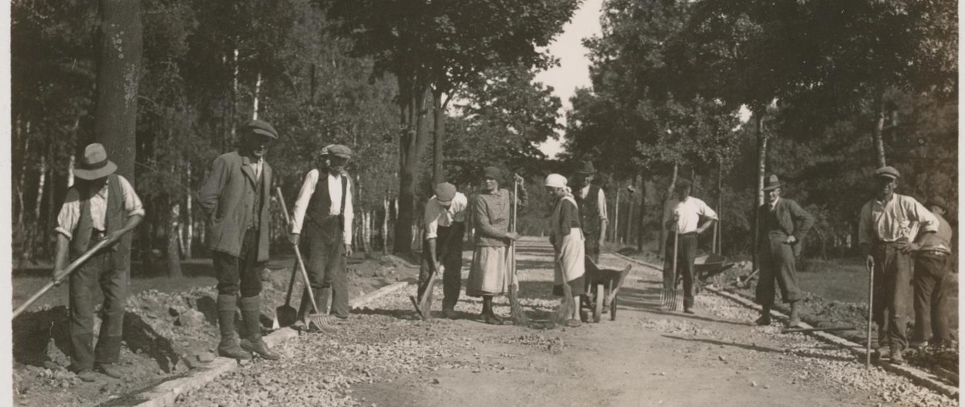 Budowa drogi w lesie, grupa ludzi trzyma proste narzędzia (łopaty, kilofy, taczki)