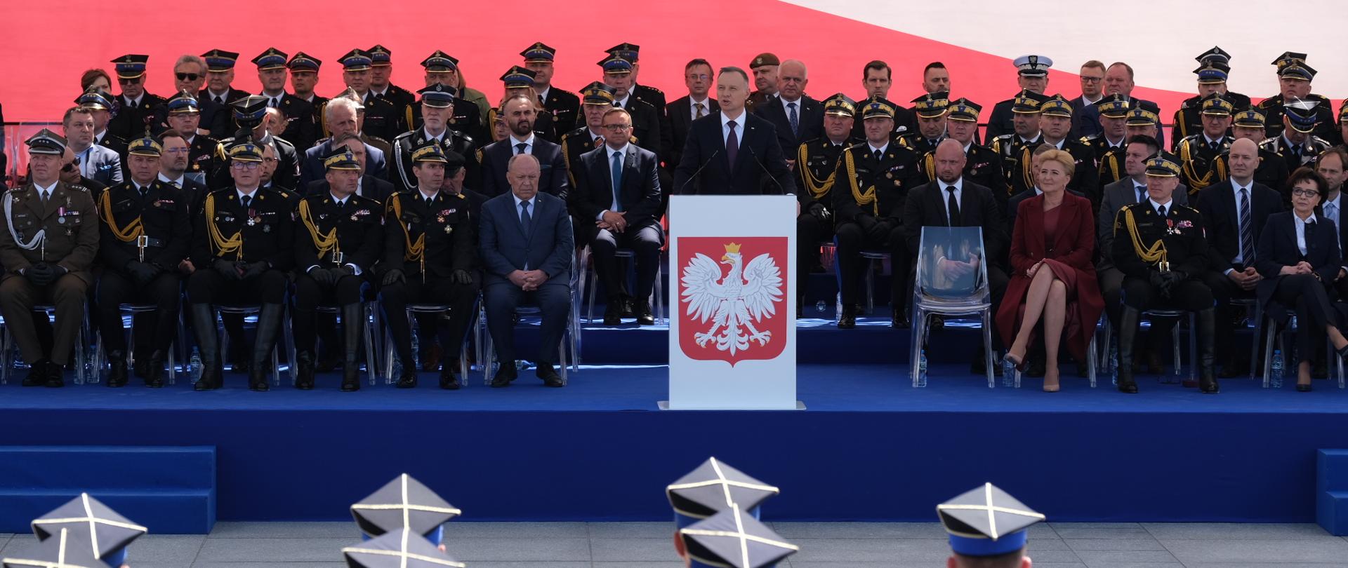 Widok z góry na strażaków stojących na placu oraz na trybunę z gośćmi, przemawia do mikrofonu prezydent RP