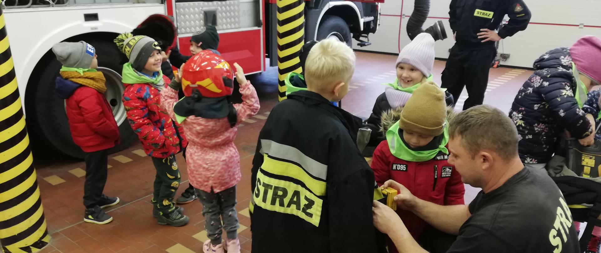 W garażu Jednostki Ratowniczo-Gaśniczej w Człuchowie dzieci z przedszkola zapoznają się ze sprzętem umieszczonym na samochodach, ubierają kurtki strażackie i hełmy.
