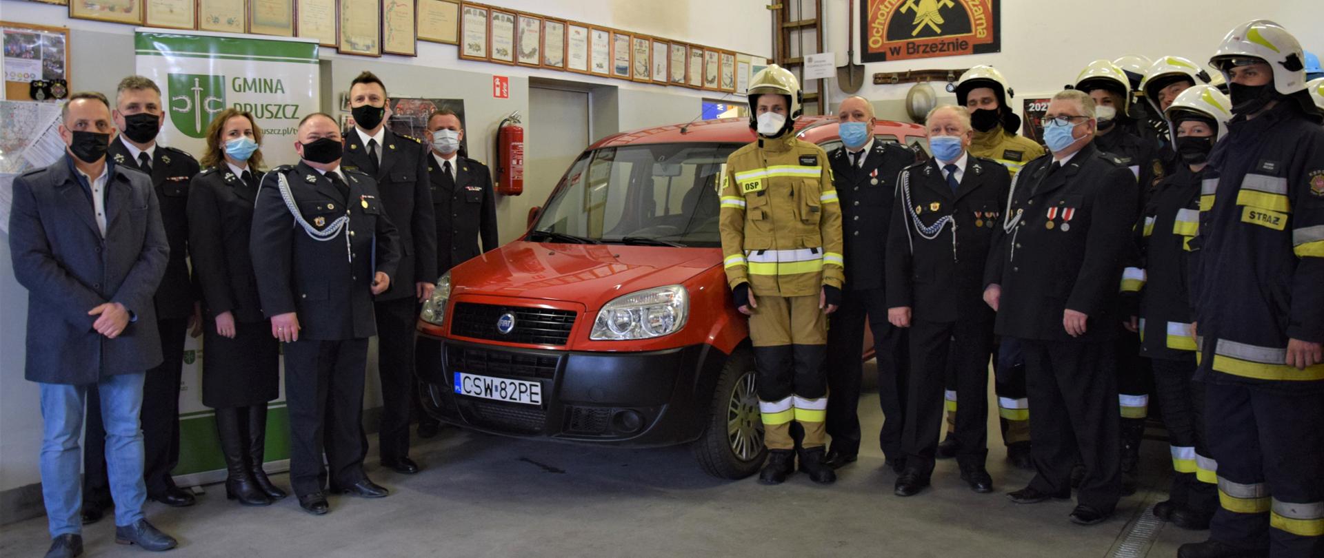 Wewnątrz garażu Ochotniczej Straży Pożarnej w Brzeźnie na środku widać przód od lewej strony samochodu strażackiego Fiat Doblo po prawej stronie samochodu stoi pięciu strażaków w mundurach wyjściowych oraz jeden cywil. Po lewej trzech strażaków w mundurach wyjściowych oraz ośmiu w mundurach specjalnych w hełmach. 