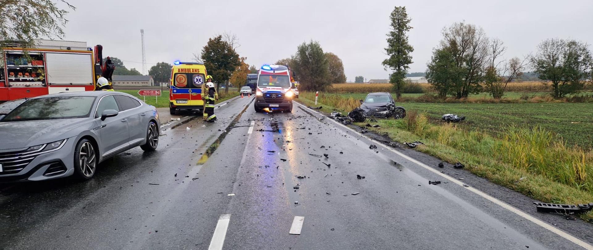 W przydrożnym rowie stoi samochód z uszkodzonym przodem. Na jezdni stoi samochód osobowy, dwie karetki pogotowia ratunkowego oraz samochód strażacki. Na jezdni porozrzucane plastiki. 