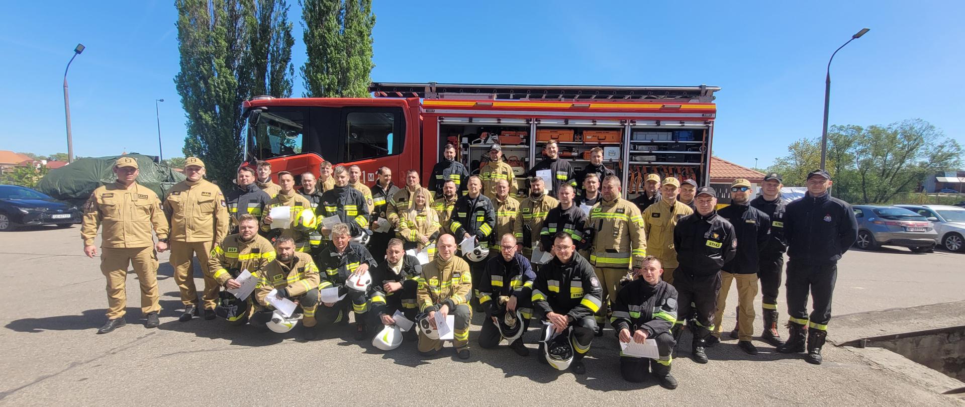 Duża grupa strażaków Ochotniczej Straży Pożarnej na tle samochodu czerwonego za nimi widać samochody osobowe i w oddali drzewa