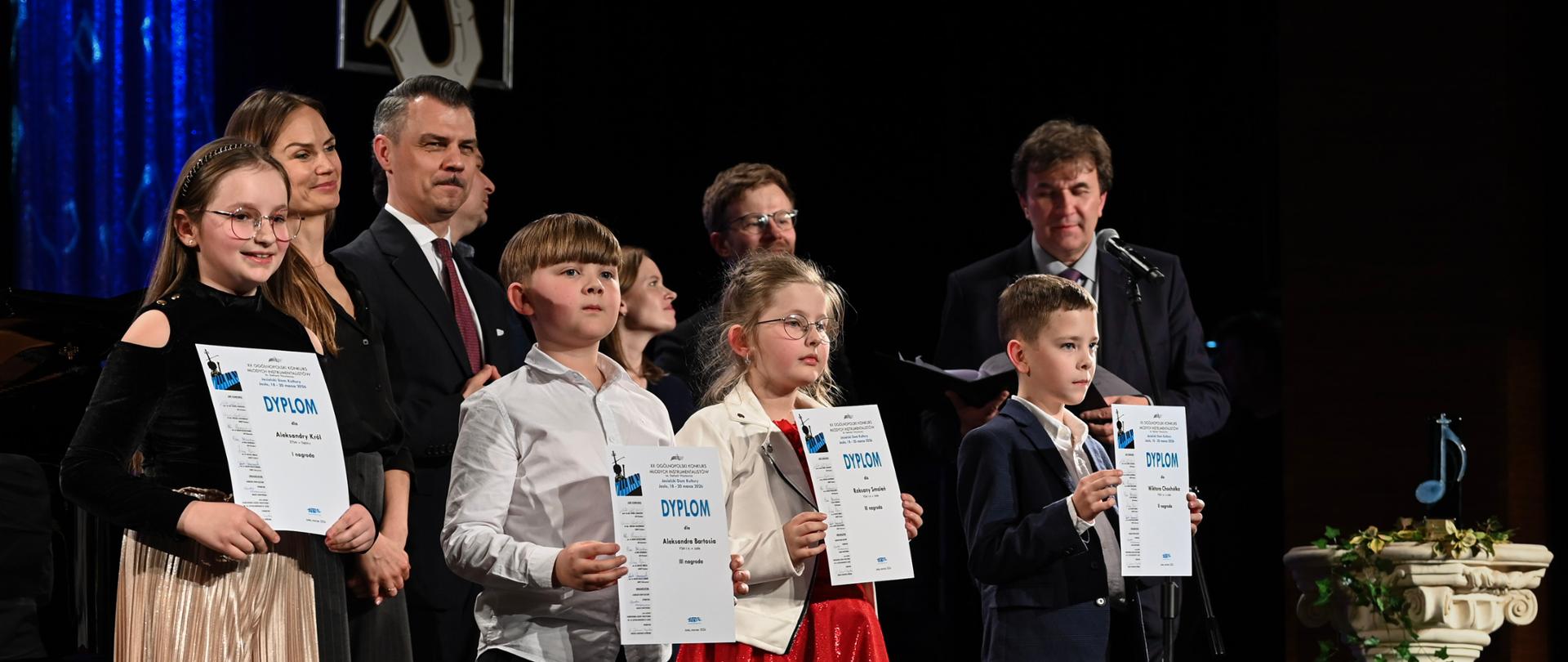 na zdjęciu, w sali widowiskowej JDK stoją nagrodzeni uczniowie, którzy trzymają w rękach dyplomy. Wraz z uczniami do zdjęcia pozują członkowie jury. Kolorystyka zdjęcia jest biało-szaro-niebiesko-brązowo-zielono-czerwono-złoto-czarna.