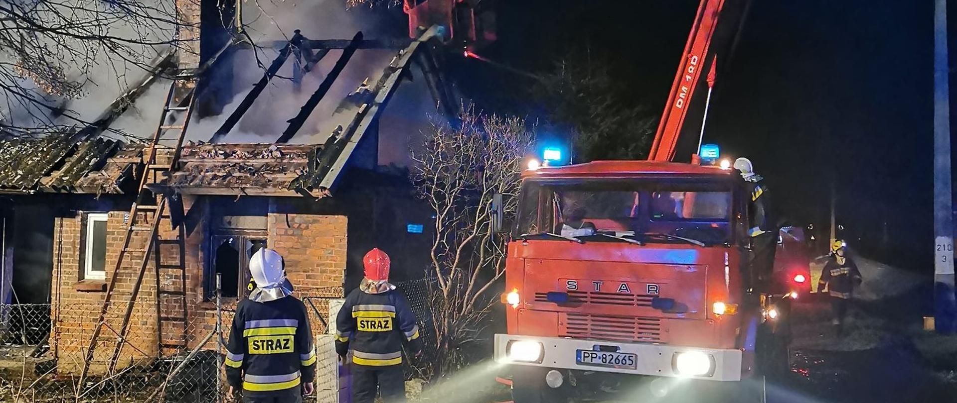 Na zdjęciu widnieją strażacy prowadzący działania gaśnicze wobec palącego się pustostanu oraz podnośnik hydrauliczny