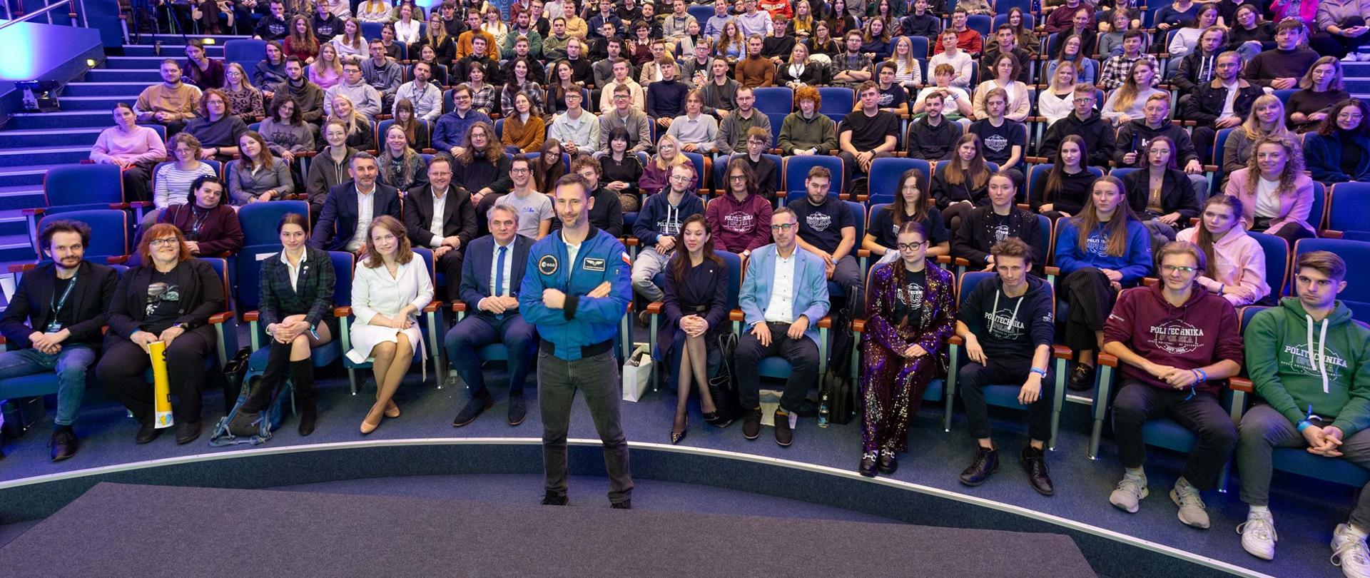 Na dużej sali w półkolistych rzędach krzeseł siedzi wiele osób, przed nimi na szarej podłodze stoi Sławosz Uznański-Wiśniewski.