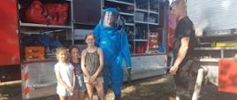 Zdjęcie przedstawia dzieci stojące przy strażakach oraz samochodzie strażackim. Jeden ze strażaków ubrany w ciężkie ubranie gazoszczelne koloru niebieskiego 