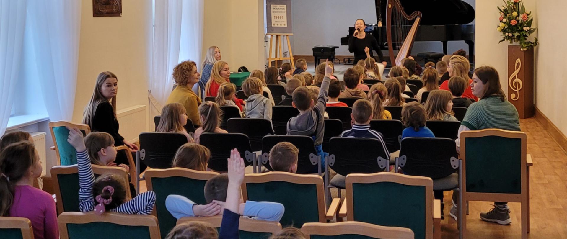 Fotografia przedstawia sylwetki dzieci - przedszkolaków wraz z paniami opiekunkami podczas koncertu "Poranek muzyczny", z przodu pani prowadząca, w oddali na scenie fortepian z otwartą klapą oraz harfa, z lewej strony sztaluga z plakatem na którym widnieje tekst Poranek muzyczny.