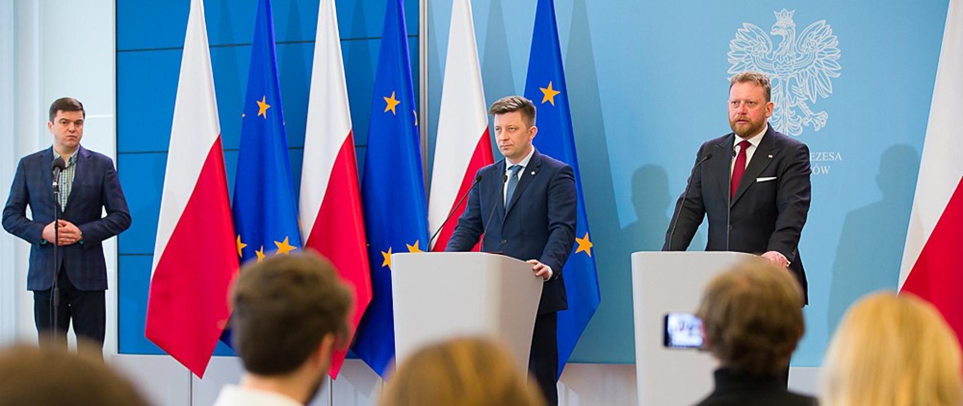 Minister Michał Dworczyk i minister Łukasz Szumowski stoją podczas konferencji prasowej.
