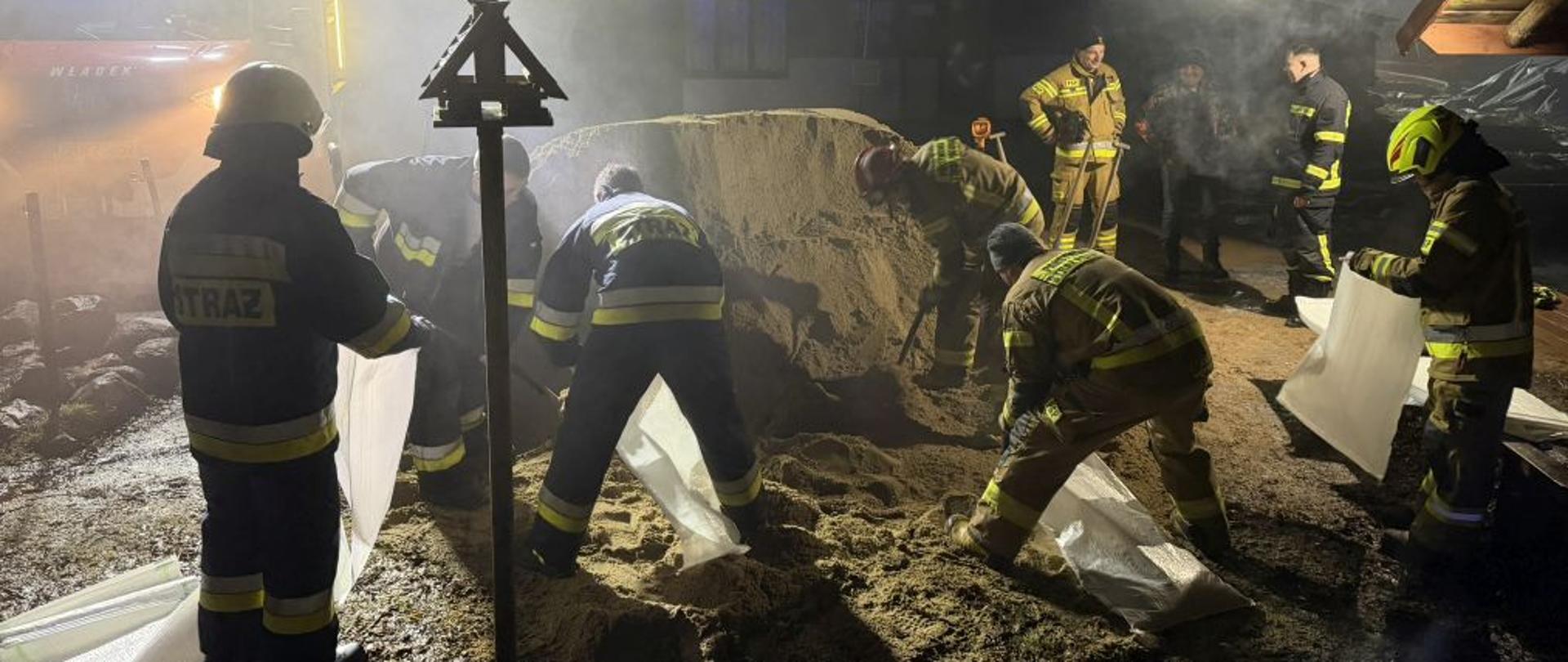 Nocne zdjęcie strażaków pracujących przy pryzmie piasku w gęstym dymie lub mgle. Pięciu strażaków pochyla się nad piaskiem, trzymając białe worki. Są ubrani w granatowe i piaskowe mundury bojowe. W tle, za unoszącymi się oparami, widać wóz strażacki marki Iveco z napisem „WŁADEK” na masce oraz włączone niebieskie światła błyskowe. Na pierwszym planie po lewej stoi drewniany słupek z daszkiem (karmnik lub drogowskaz).