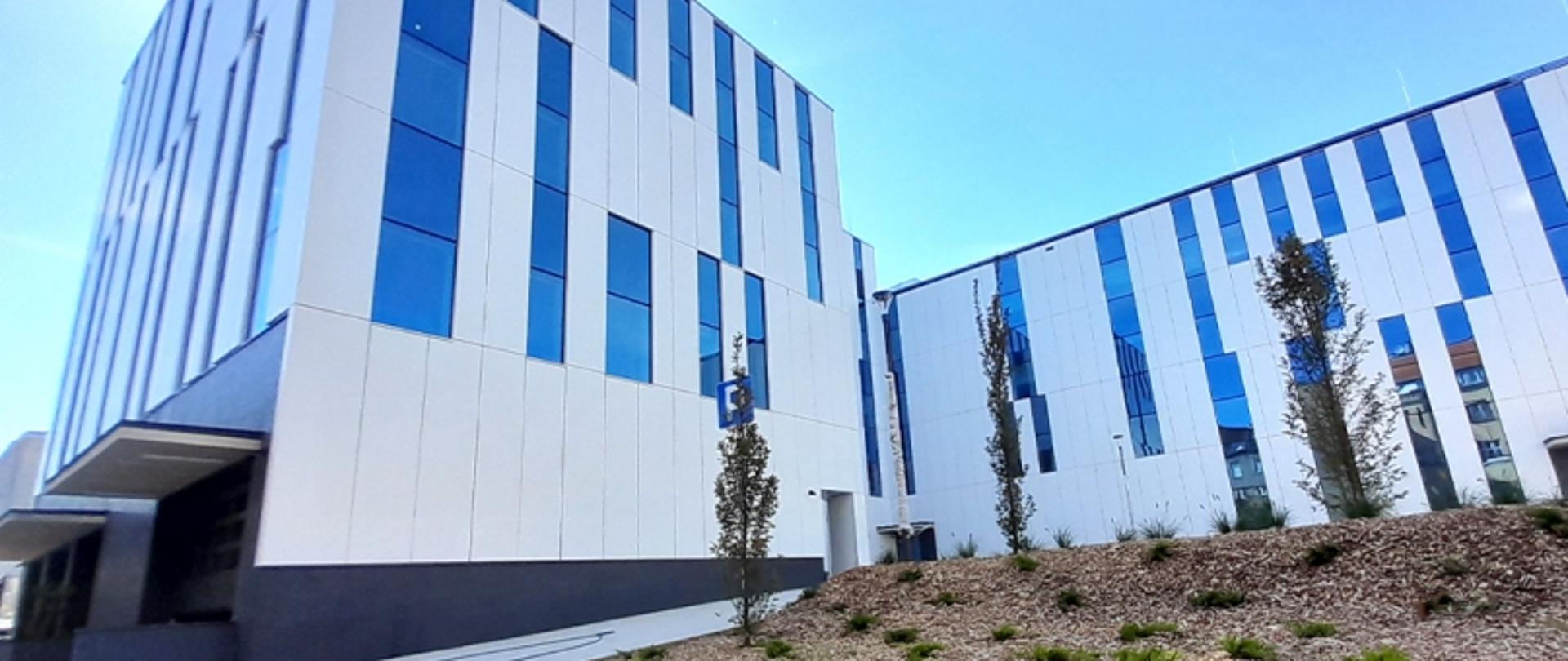 Dwa nowoczesne budynki z pionowymi panelami w kolorach białym i niebieskim, otoczone roślinnością, pod błękitnym niebem.