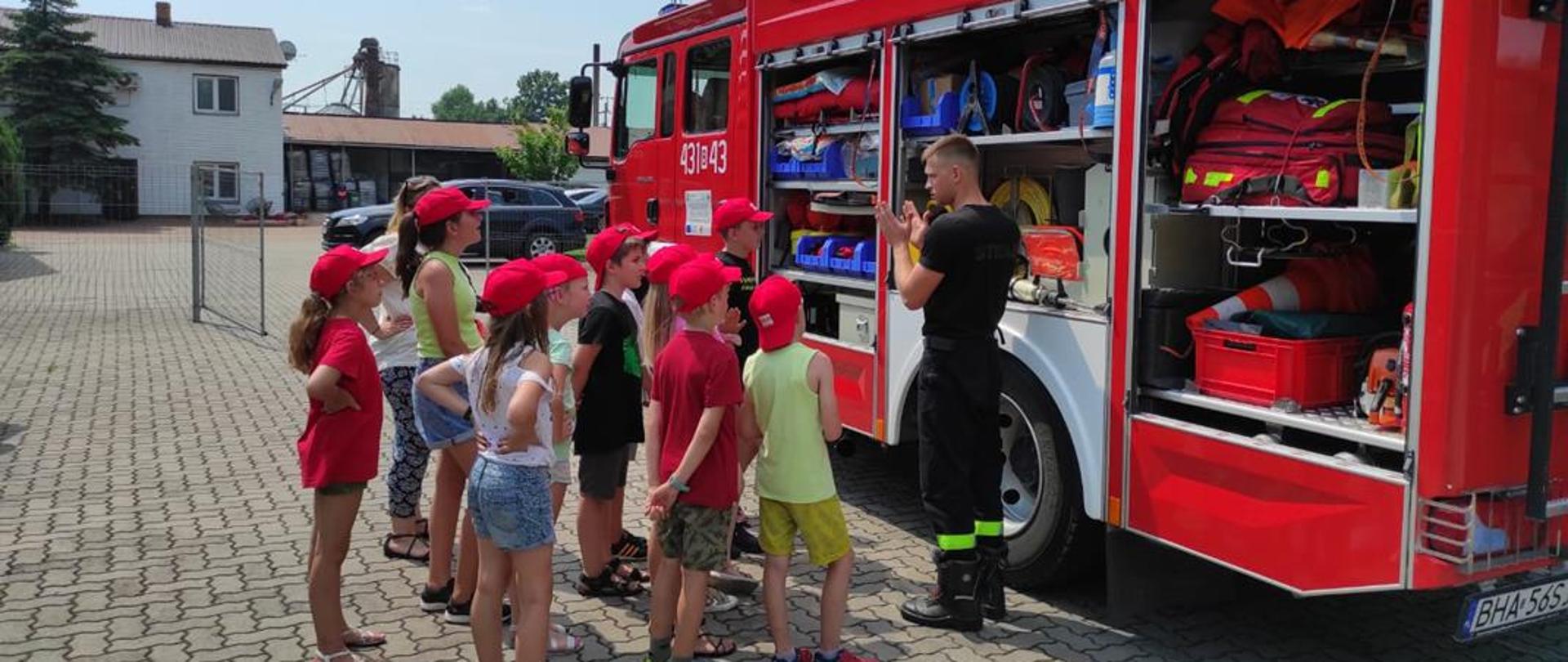 Strażak stojący przy samochodzie pożarniczym pokazuje dzieciom jego wyposażenie.