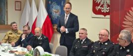 Przy stołach na których znajdują się dekoracje świąteczne i talerze, siedzą strażacy i zaproszenie goście. Strażacy ubrani są w mundury galowe zaproszeni goście odświętnie w garniturach. Prezydent miasta w postawie stojącej przemawia. W tle logo Państwowej Straży Pożarnej oraz godło Polski i flagi