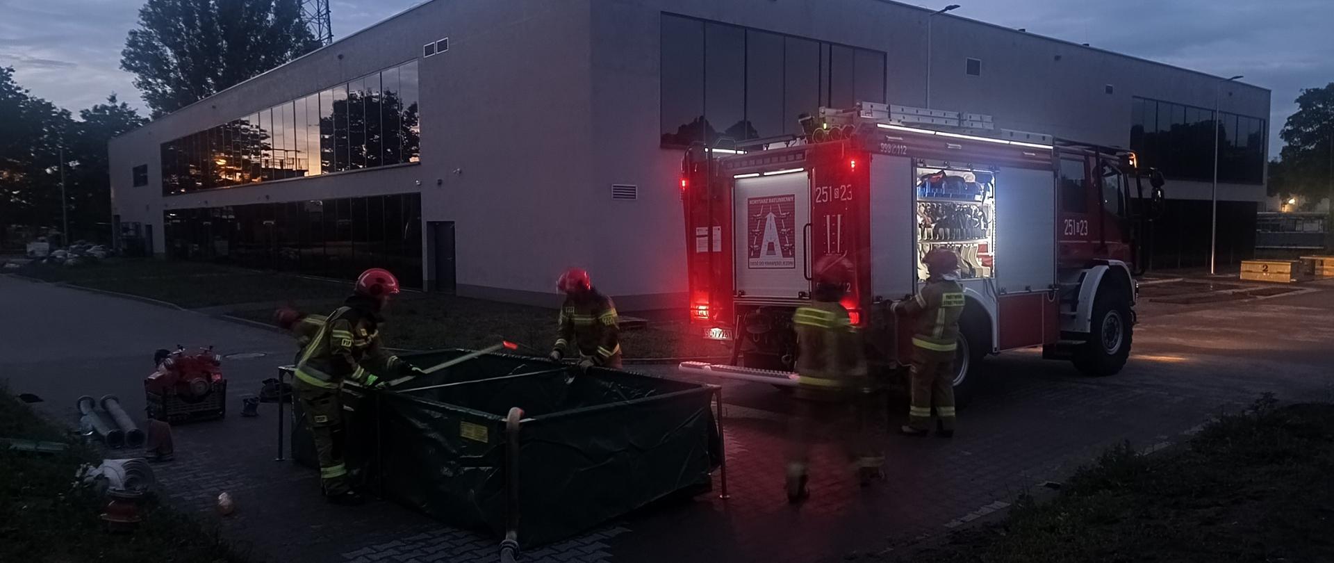 Poligon Szkolny wieczorową porą, dwóch strażaków działa przy zbiorniku z wodą rozstawionym na drodze dojazdowej, dwóch kolejnych znajduje się przy wozie bojowym stojącym po prawej stronie. W tle budynek hali sportowo-laboratoryjnej