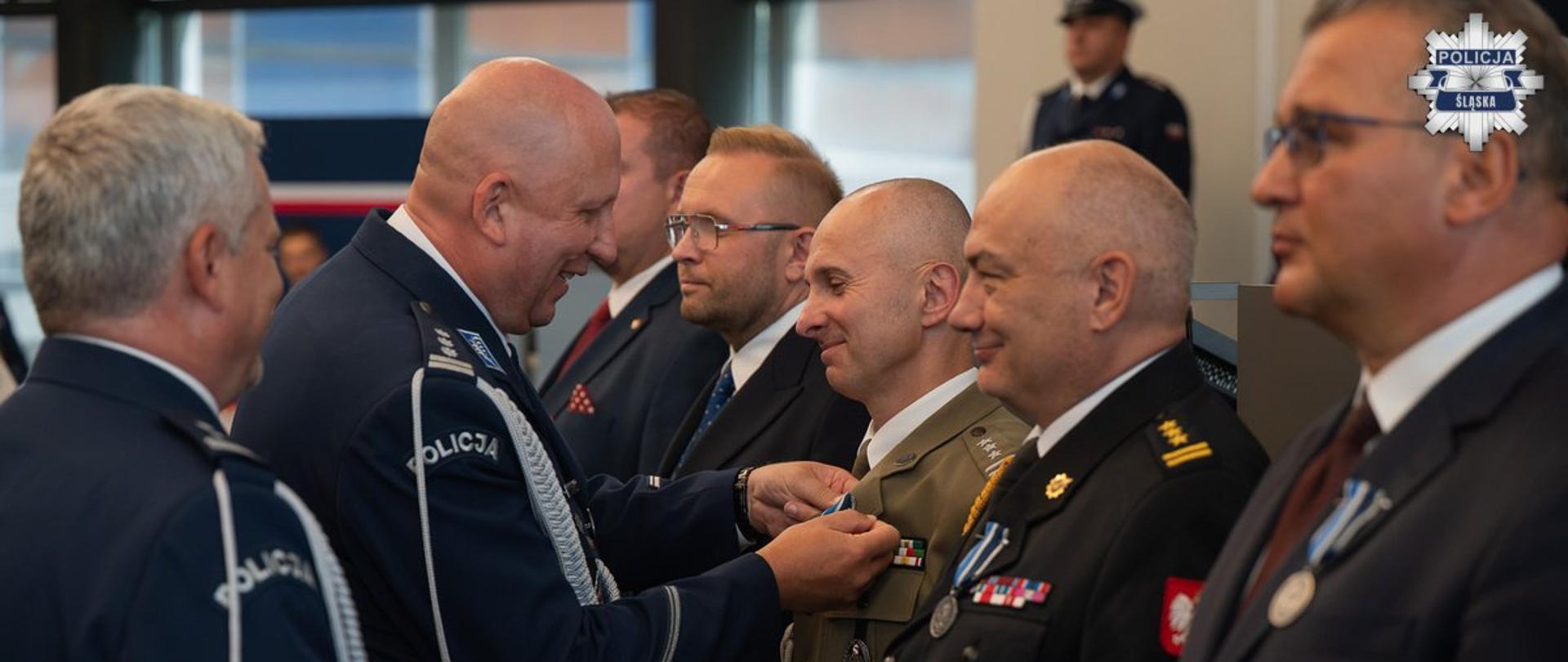 Inspektor Policji przypina medal do klapy munduru pułkownika Wojska Polskiego, stojącego w rzędzie wraz z innymi odznaczanymi osobami
