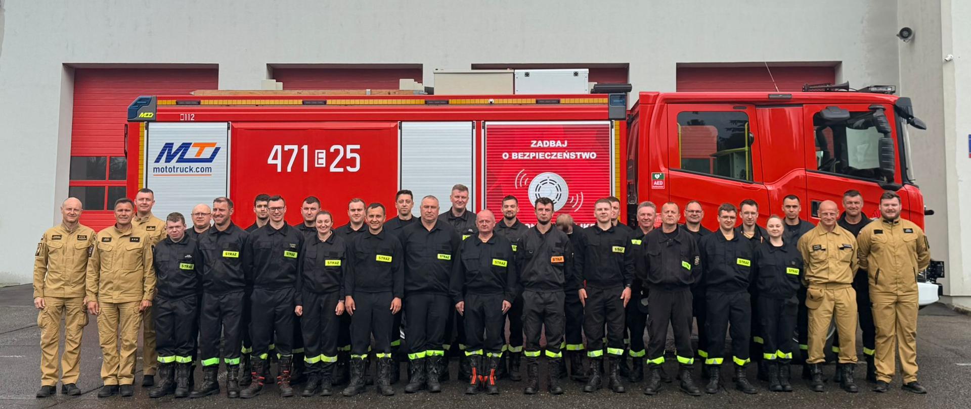 Grupa strażaków w ubraniach koszarowych koloru czarnego i piaskowego. Trzech funkcjonariuszy w ubraniach służbowych koloru piaskowego na tle samochodu pożarniczego i budynku Komendy Powiatowej. 