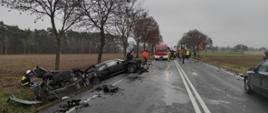 Dwa samochody osobowe w rowie po zderzeniu czołowym. Jeden samochód na boku. Na jezdni porozrzucane fragmenty karoserii. Na dalszym planie wóz strażacki i strażacy.
