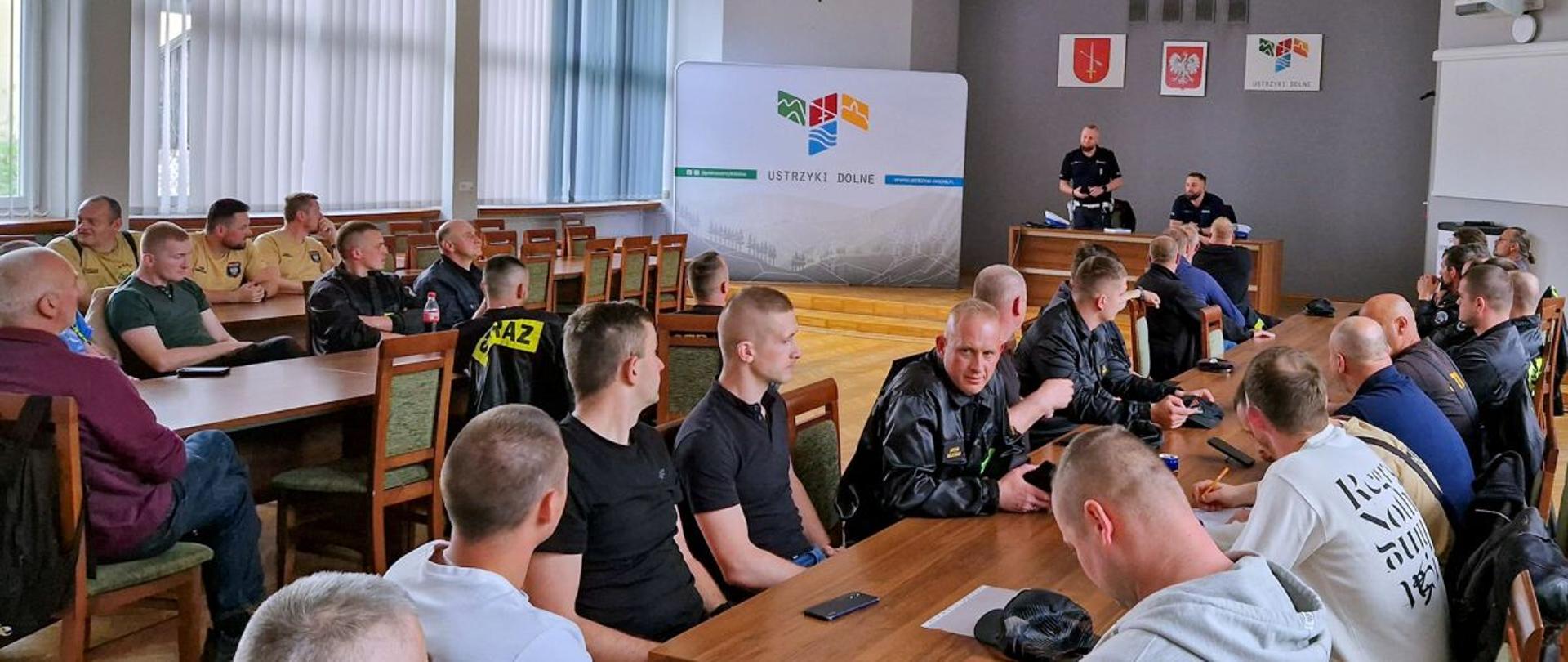 Zdjęcie przedstawia strażaków Państwowej Straży Pożarnej i Ochotniczych Straży Pożarnych z powiatu Bieszczadzkiego, część ubrana jest w ubrania koszarowe a część w ubrania cywilne. Przy biurku pod ścianą jeden Policjant siedzi a jeden stoi. Zdjęcie wykonano w sali urzędu miasta. Na ścianie zawieszone jest Godło Polski, Herb i logo Ustrzyk Dolnych. 