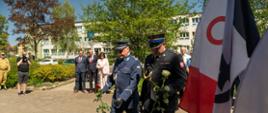 Fotografia przedstawia oficjalny moment składania kwiatów podczas uroczystości o charakterze patriotycznym, odbywającej się w słoneczny dzień na świeżym powietrzu.