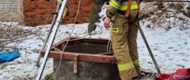 Strażak ubrany w szelki ratownicze przy studni gdzie wpadł pies, nad dryną studni rozstawiony trójnóg ratowniczy, pora dzienna.