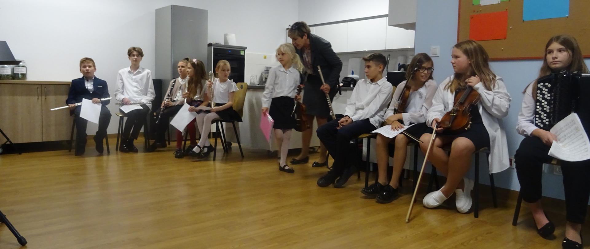 Zdjęcie siedzących uczniów w sali przed koncertem, trzymają instrumenty.