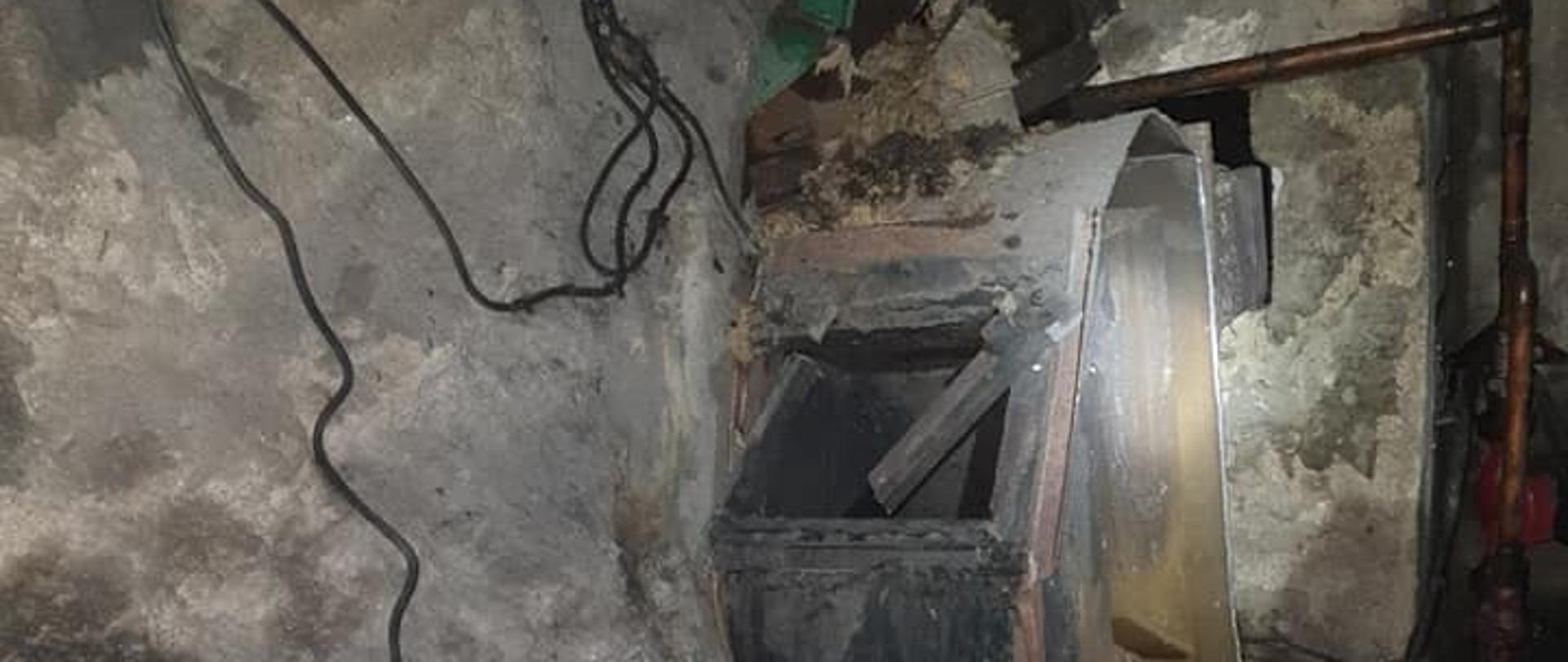 Na zdjęciu przedstawiony jest piec który uległ uszkodzeniu podczas jego użytkowania. Znajduję się w kotłowni w części podpiwniczonej budynku wielorodzinnego. Ściany wokół są uszkodzone, wyglądają jak po pożarze.