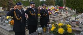 Widok z przodu. Na cmentarzu przed grobem stoją trzej strażacy w mundurach wyjściowych i salutują. 