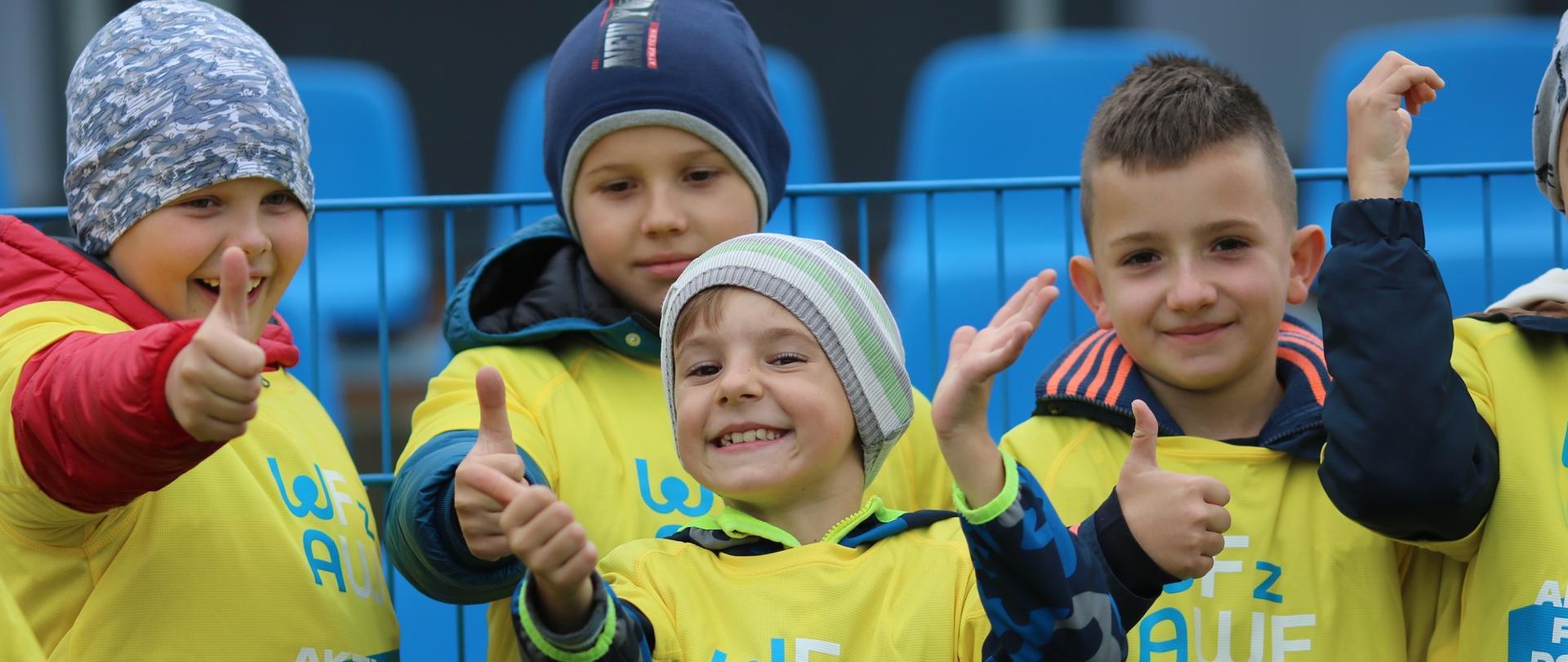 Grupa wesołych ubranych na zielono dzieci stoi na boisku, pokazują kciuki w górę.