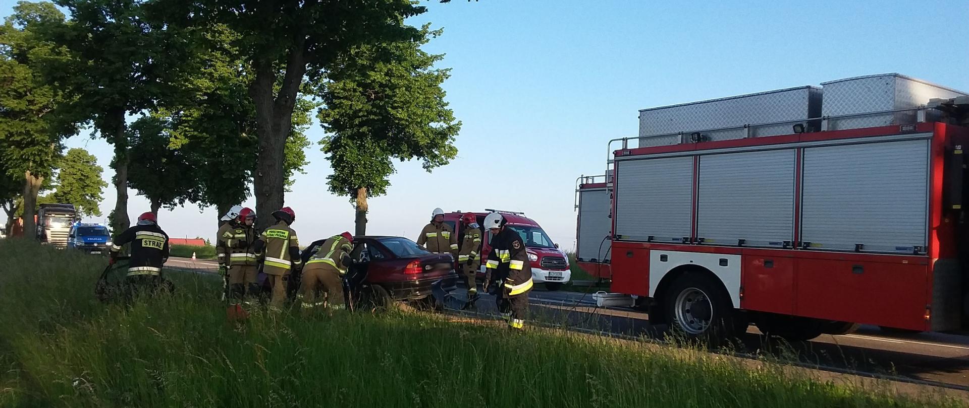Ujęcie z pobocza drogi, w centrum kadru w oddali widoczny rozbity na drzewie samochód osobowy i pracujący strażacy. Na jezdni stoją pojazdy pożarnicze. 