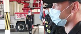 Dowódca uroczystości - starszy kapitan Damian Łojek podaje rozkaz do wyprowadzenia pocztu sztandarowego. Z prawej strony zdjęcia widoczni w szeregu strażacy Jednostki Ratowniczo-Gaśniczej. W tle widoczny czerwony samochód pożarniczy.