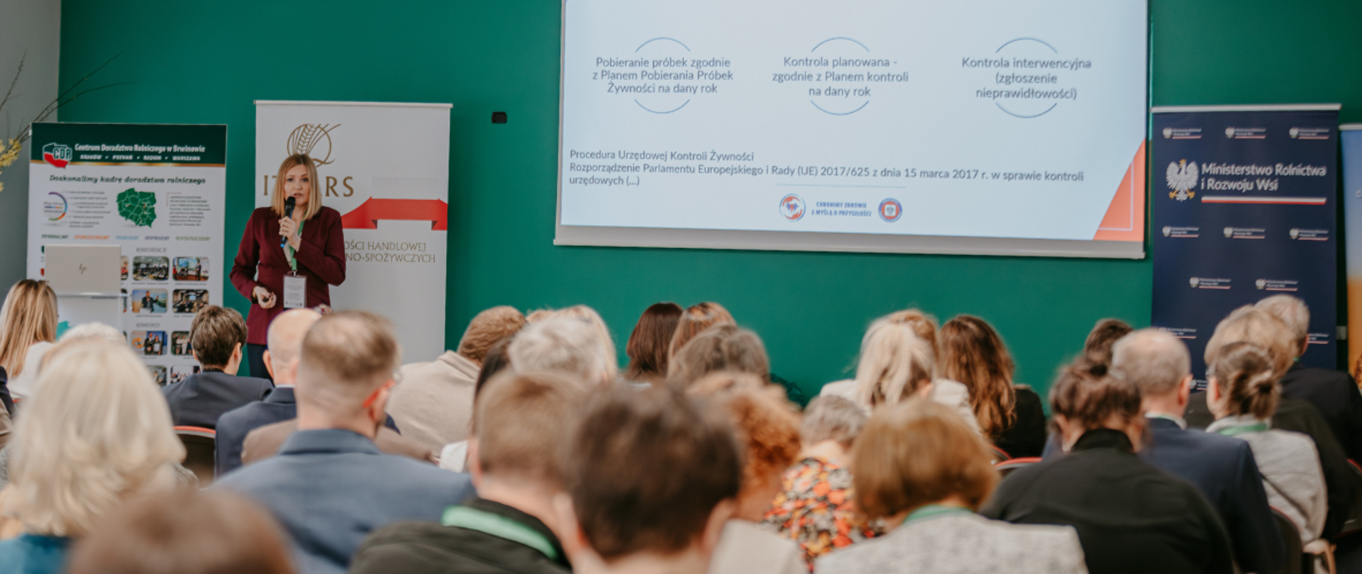 Sala konferencyjna, uczestnicy siedzą tyłem na krzesłach. W centralnej części ekran z prezentacją przedstawicielki WSSE. Prowadząca stoi po lewej stronie. 