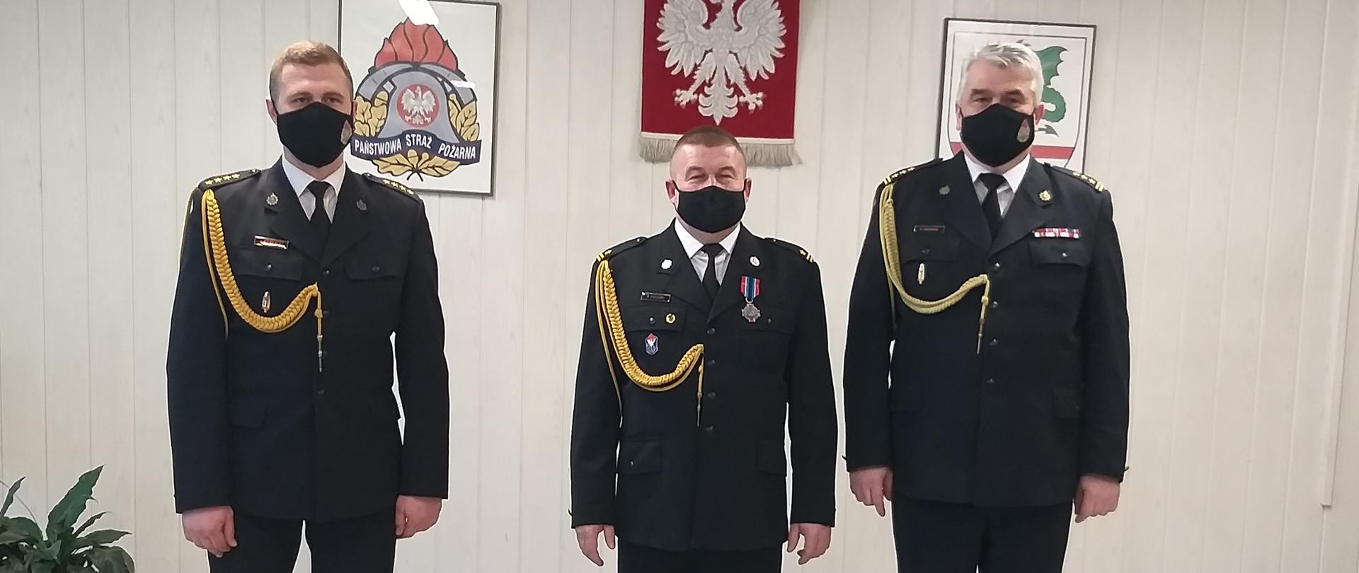 Trzech funkcjonariuszy ubranych w mundur galowy stojących w sali konferencyjnej. W tle na ścianie wisi godło Rzeczypospolitej. logo PSP oraz goło starostwa.
