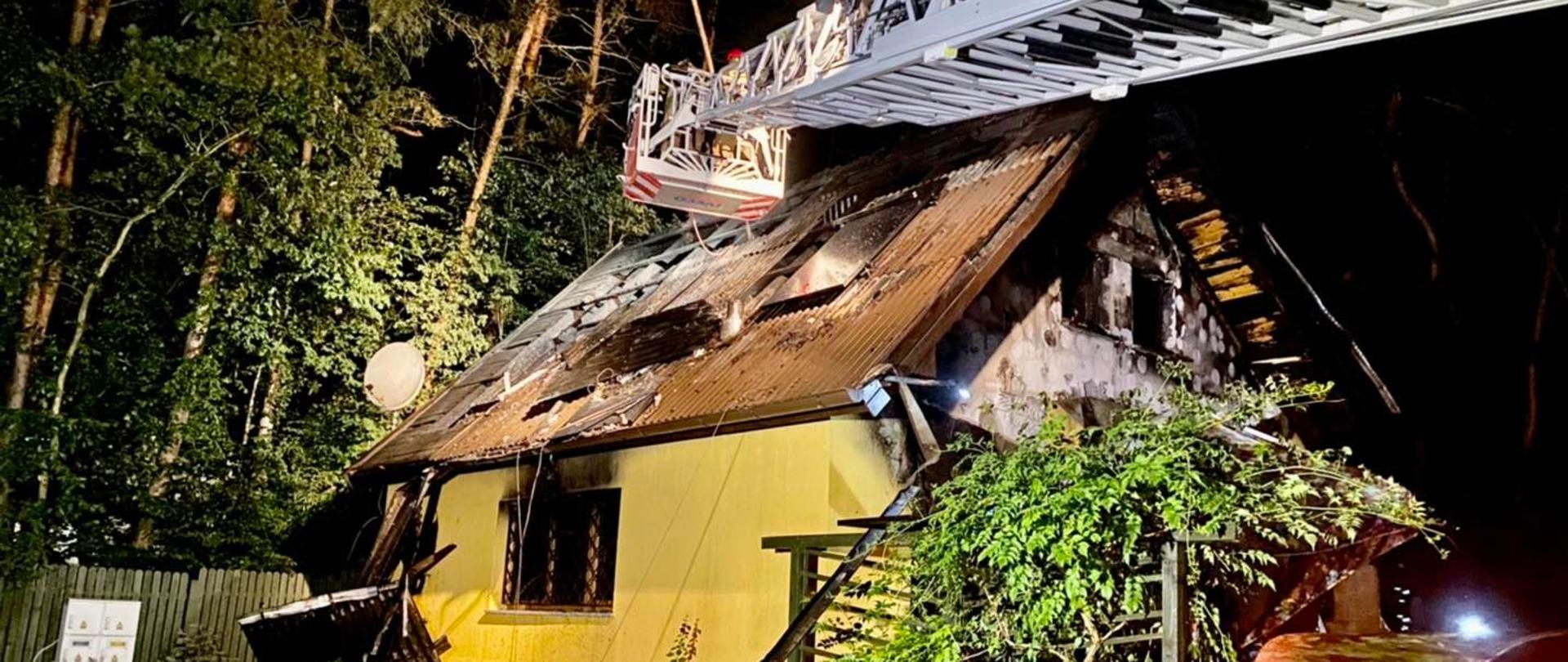 Pora nocna. Budynek letniskowy oświetlony sztucznym światłem z częściowo spalonym dachem i elewacją . Nad nim metalowe przęsło i kosz drabiny mechanicznej. Obok niego spalone panel fotowoltaiczne