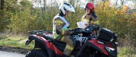 Widok z boku. Kierujący działaniem ratowniczym siedzi na quadzie w białym kasku i przyjmuje meldunek od dowódcy odcinka bojowego - funkcjonariusza PSP. 