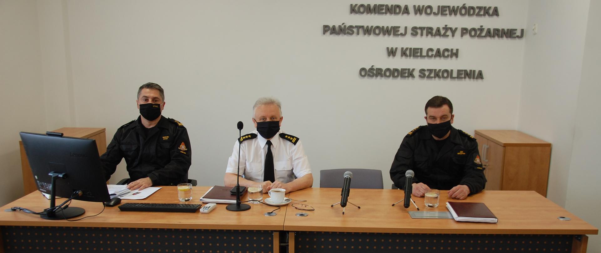 Zdjęcie przedstawia uczestników spotkania siedzących za stołem prezydialnym to jest: Zastępcę Świętokrzyskiego Komendanta Wojewódzkiego PSP oraz naczelników Ośrodka Szkolenia i Wydziału Operacyjnego KW PSP w Kielcach w umundurowaniu służbowym.
