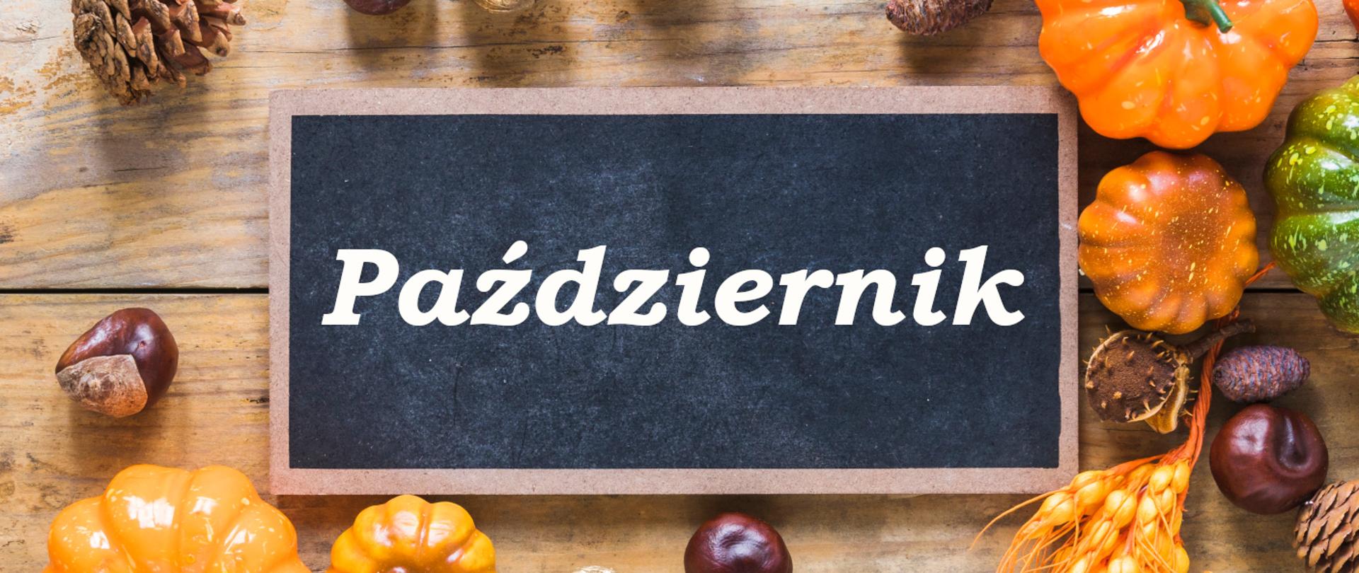 Ciemna tabliczka z białym napisem październik na drewnianym stole w otoczeniu kasztanów, szyszek i dyń. 