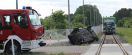 Samochód leżący na boku przy nasypie kolejowym po zderzeniu z szynobusem. Przed pojazdem samochód ratowniczo-gaśniczy. W tle szynobus.
