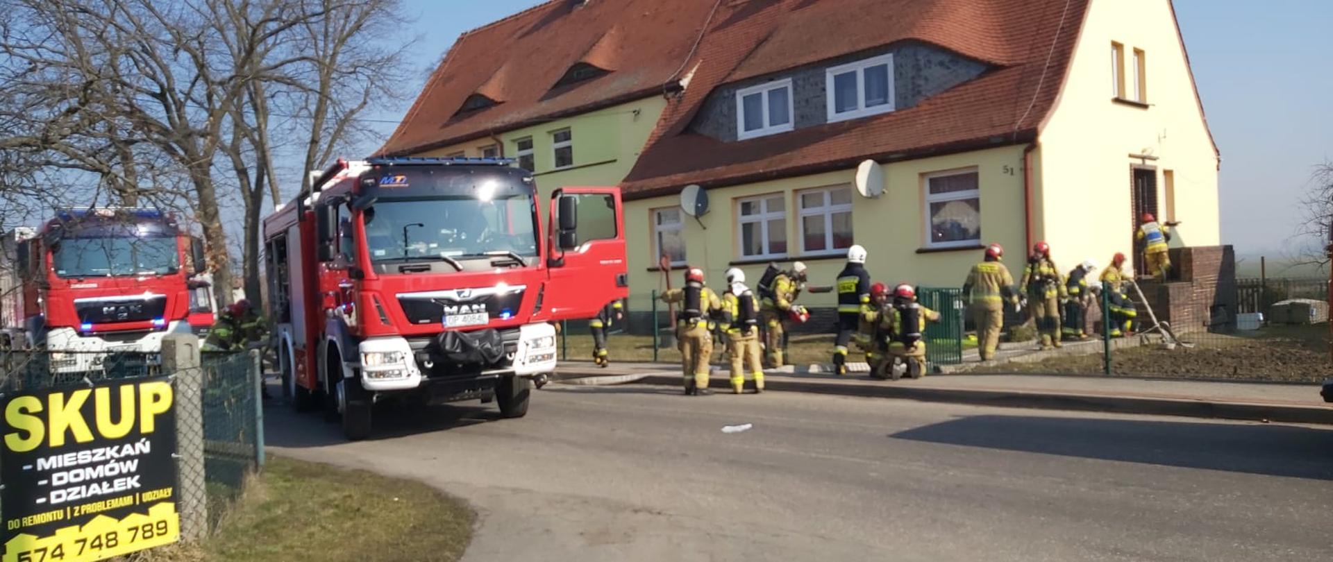 Zdjęcie przedstawia wstępny etap organizacji akcji gaśniczej. Na tle budynku wielofunkcyjnego w kolorze bladożółtym z dachem z pomarańczowej dachówki, po lewej stronie widać dwa samochody pożarnicze. Przy nich znajdują się strażacy wyjmujący sprzęt niezbędny do działań. W środku zdjęcia znajdują się strażacy PSP i OSP przygotowujący się do wejścia do obiektu i rozwijający węże linii gaśniczych. Strażacy PSP w umundurowaniu specjalnym koloru piaskowego i czerwonych hełmach. Ratownicy OSP w mundurach piaskowych lub granatowych w hełmach białych. Po prawej stronie budynku widać wejście na klatkę schodową do której wchodzi dowódca na rozpoznanie. 