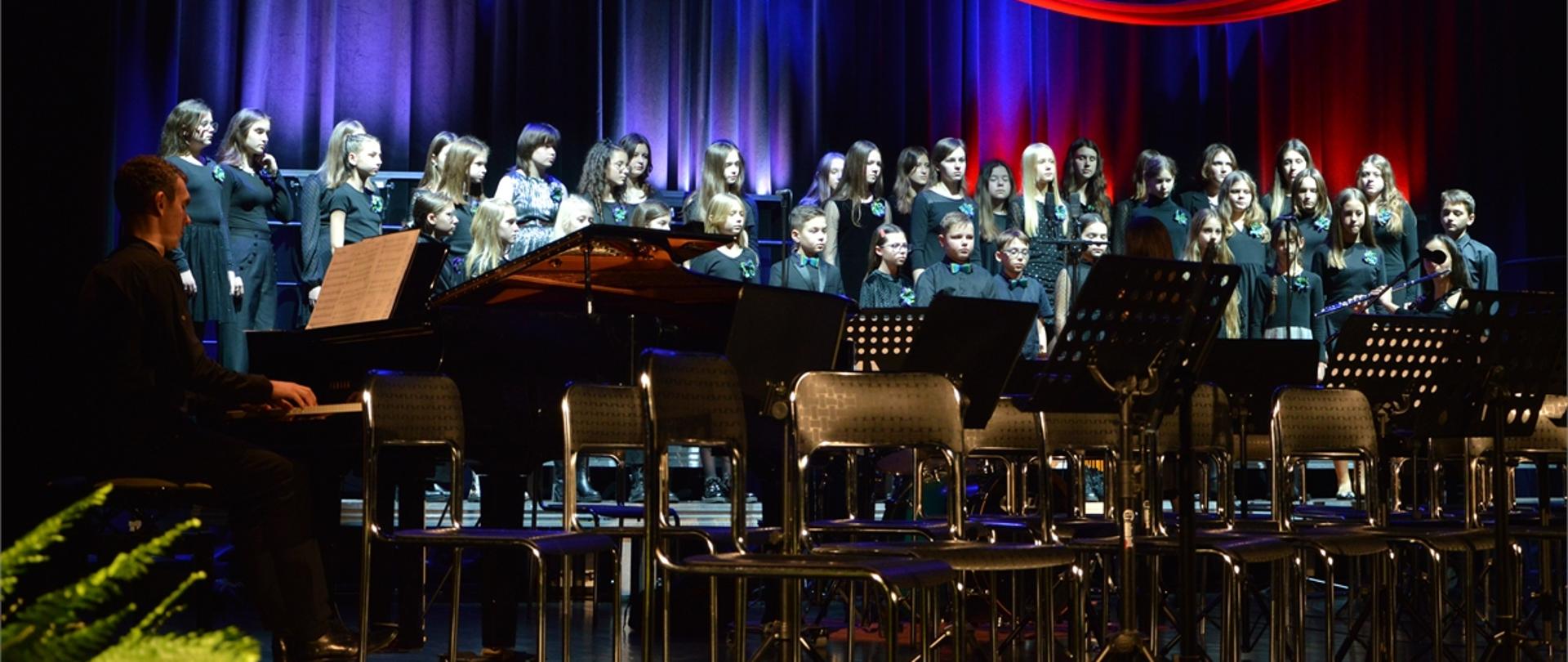 Zdjęcie przedstawia chór szkolny występujący na scenie Jasielskiego Domu Kultury, w tle biało-czerwona wstęga, kolorystyka zdjęcia niebiesko-czarna.