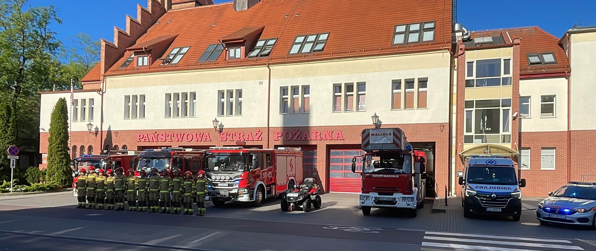 Strażnica KM PSP Sopot, przed którą wystawione sa pojazdy PSP, OSP oraz dwie karetki. Przed samochodami stoja strażacy w ubraniach specjalnych i hełmach, ustawieni na baczność w jednym szeregu oddając hołd strażakowi KG który zginął