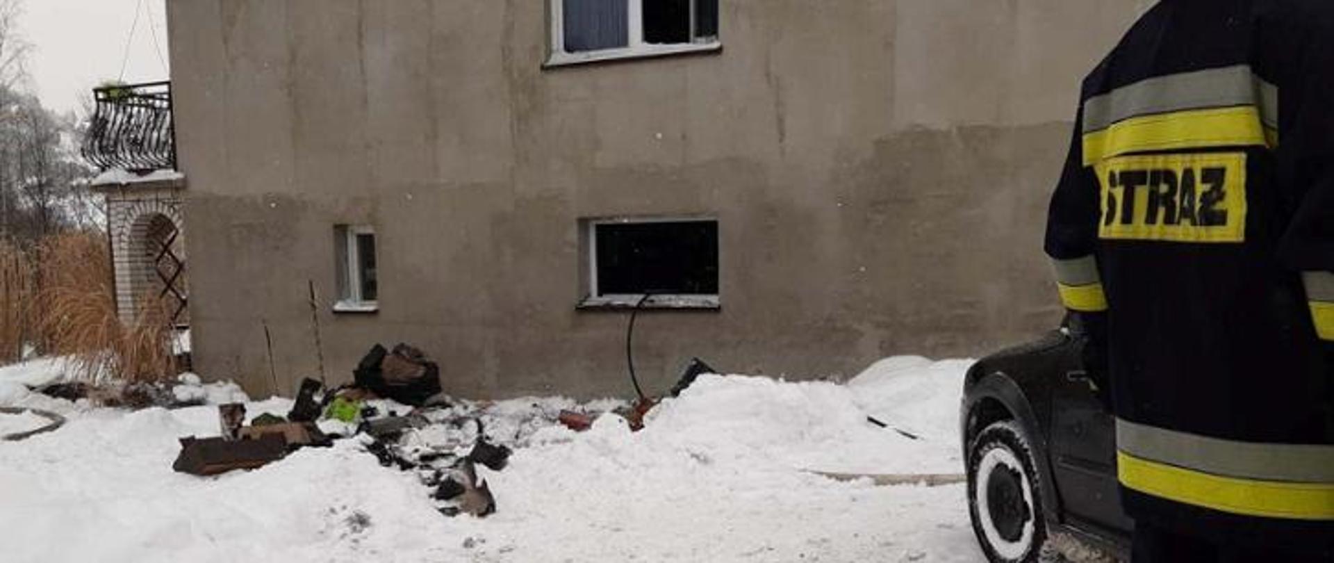 Budynek mieszkalny, wokół śnieg. Z lewej strony widać tyłem stojącego strażaka.
