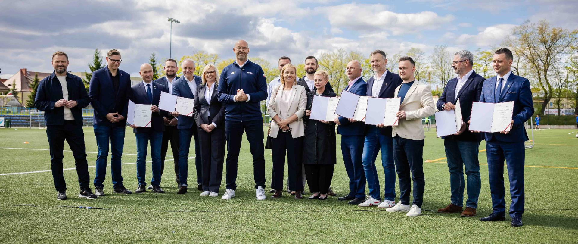 Zdjęcie zbiorowe na boisku piłkarskim. Minister sportu i turystyki Jakub Rutnicki i przedstawiciele lokalnych władz prezentują decyzje ministra dotyczące dofinansowań dla pełnowymiarowych boisk piłkarskich w województwie zachodniopomorskim.