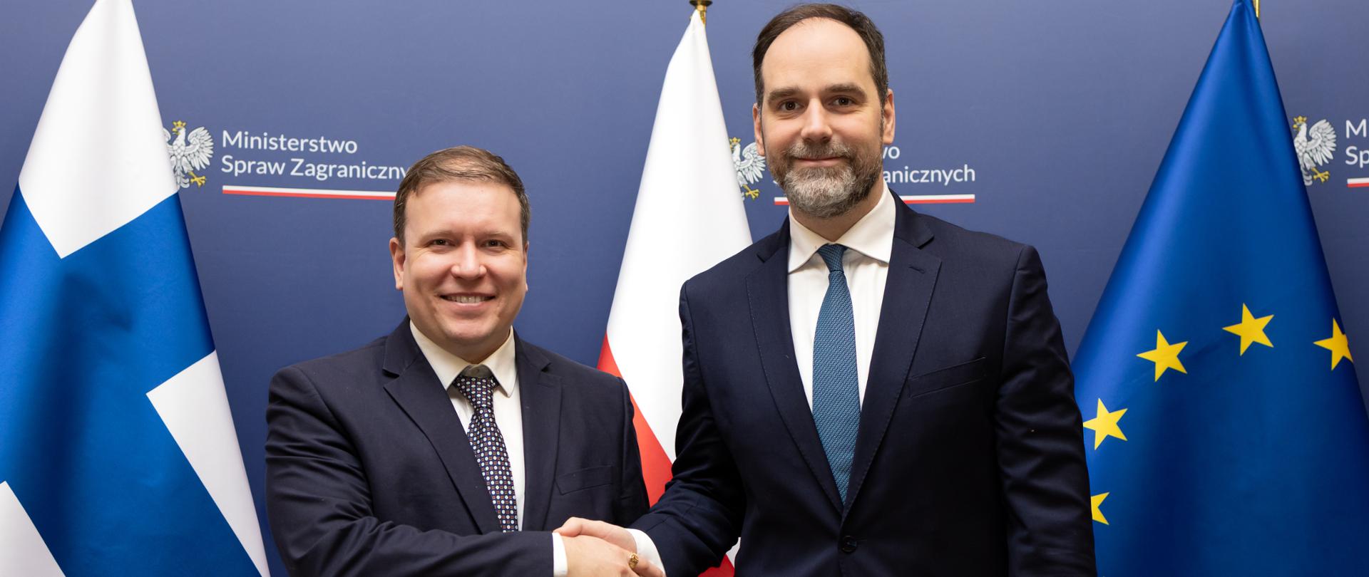Dwóch mężczyzn stoi na tle flag narodowych i flagi Unii Europejskiej, podając sobie dłonie. Zdjęcie wykonane w przestrzeni instytucjonalnej, z widocznym oznakowaniem ministerstwa. Ujęcie ma charakter oficjalny i protokolarny.