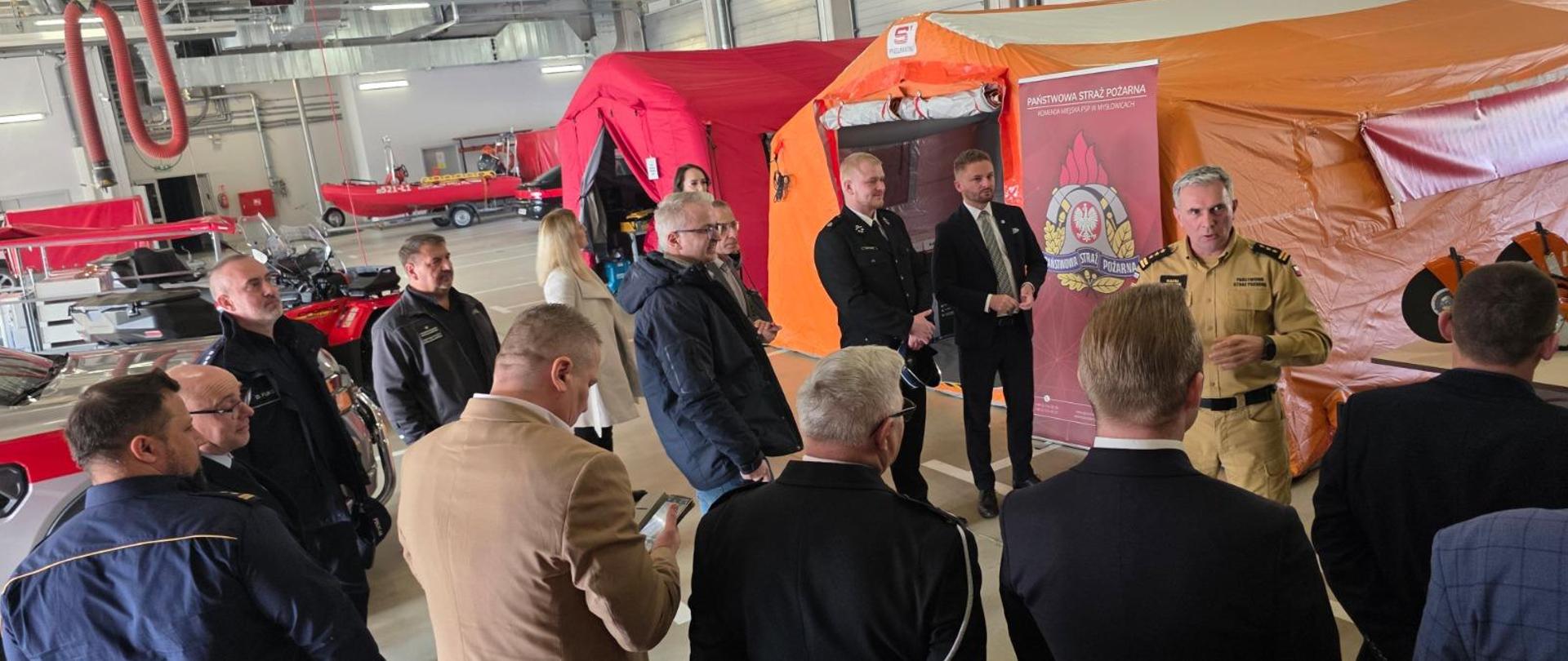 Następne zdjęcie dokumentuje wizytację zaproszonych gości w hali garażowej, gdzie w praktyczny sposób prezentowane jest wyposażenie jednostki.
Oto szczegółowy opis tej części wydarzenia:
Prezentacja Sprzętu i Rozmowy
Prowadzący: Oficer w piaskowym mundurze służbowym (widoczny po prawej stronie) aktywnie gestykuluje, omawiając specyfikę pracy lub parametry techniczne prezentowanego sprzętu.
Goście i Uczestnicy: Wokół prelegenta zgromadziła się grupa osób, w tym cywile w garniturach i kurtkach oraz funkcjonariusze w różnych typach umundurowania (m.in. ciemne mundury służbowe). Uczestnicy z zaciekawieniem słuchają wyjaśnień, a jeden z nich dokumentuje prezentację telefonem.
Eksponowane Wyposażenie w Tle
Sceneria hali garażowej pozwala dostrzec różnorodność zasobów KM PSP w Mysłowicach:
Namioty pneumatyczne: Bezpośrednio za grupą rozstawione są duże namioty ratownicze – pomarańczowy (z widocznym logo producenta "S-Pneumatic") oraz czerwony. Służą one jako szpitale polowe lub bazy logistyczne podczas długotrwałych akcji.
Sprzęt mechaniczny: Po prawej stronie, na stole, widoczne są tarcze przecinarek spalinowych, a po lewej fragmenty pojazdów ratowniczych oraz quad.
Ratownictwo wodne: W głębi hali widoczna jest czerwona łódź ratunkowa na przyczepie o numerze operacyjnym 521-L1.
Detale Organizacyjne
Obok grupy stoi bordowy roll-up Państwowej Straży Pożarnej, który towarzyszy wszystkim etapom narady, spójnie identyfikując miejsce wydarzenia. W górnej części hali widoczne są instalacje techniczne, w tym systemy odciągu spalin (czerwone rury) oraz nowoczesne oświetlenie.