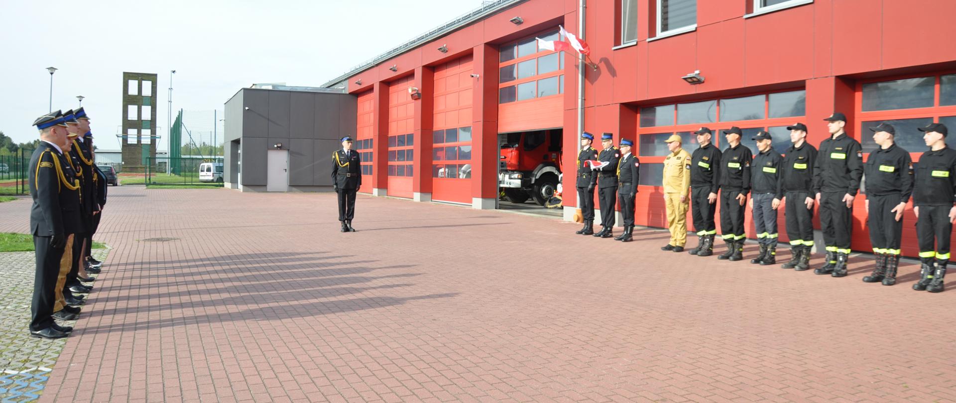 Plac przed komendą państwowej straży pożarnej w brzezinach, po prawej oraz po lewej strażacy komendy w umundurowaniu galowym, po środku prowadzący uroczystość wraz z pocztem sztandarowym gotowym do wciągnięcia flagi na maszt